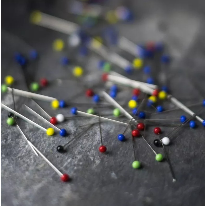 A close-up, shallow depth-of-field shot of colorful sewing pins scattered on a dark gray textured surface.
