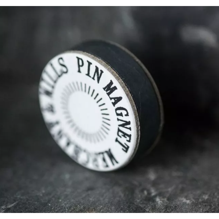 A circular black and white pin magnet tool with the text 'KILLS PIN MAGNET' printed on it, resting on a dark textured surface.