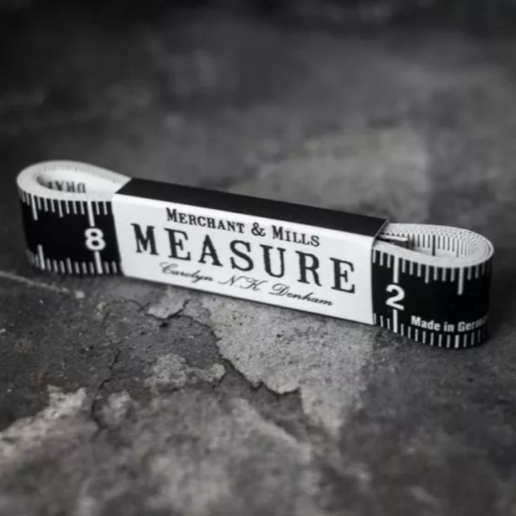 A vintage-style measuring tape coiled horizontally on a textured dark gray surface. The tape is black and white with printed measurement markings and a central paper band reading 'MERCHANT & MILLS MEASURE' and 'Carlsberg N.