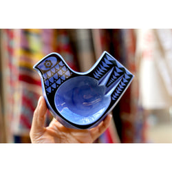 A photograph of a hand holding a blue dish in the shape of a bird with black details. In the background there are colourful fabrics.