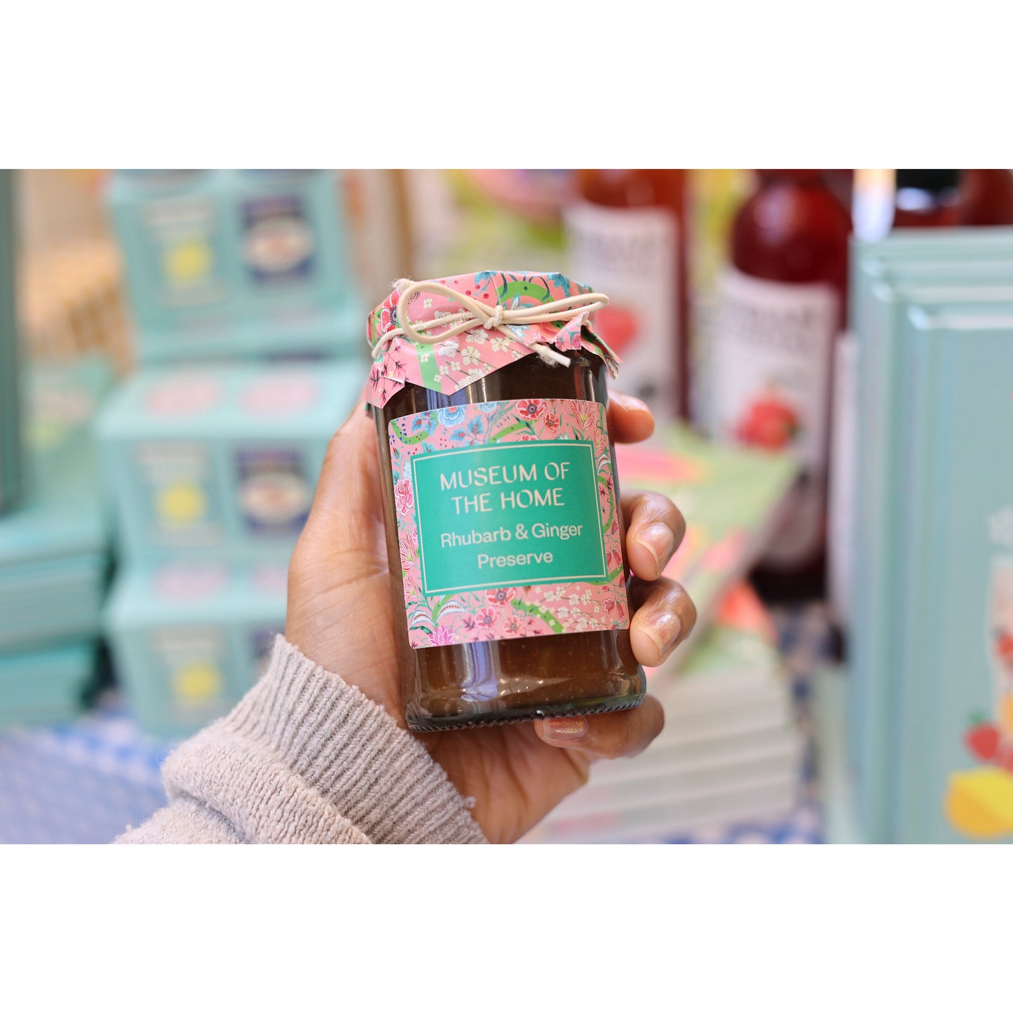 A hand holding a preserve of rhubarb and ginger. The glass jar features a pink label with a pink paper lid wrapped in ribbon. In the background it can be seen items from the shop.
