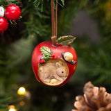 A bauble of a red apple with a mouse inside it and a ladybug and a snail on it hanging on a Christmas tree.