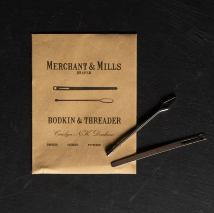 A vintage-style booklet or pattern card titled 'Merchant & Mills Draper Bodkin & Threader' lies on a dark textured surface, with two metal thread tools placed on top.