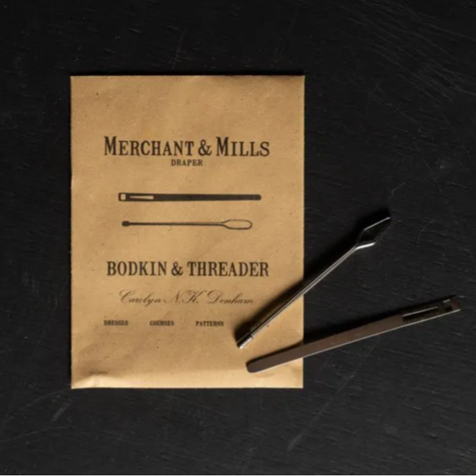 A vintage-style booklet or pattern card titled 'Merchant & Mills Draper Bodkin & Threader' lies on a dark textured surface, with two metal thread tools placed on top.
