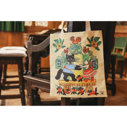 A bag hanging on a wooden chair. The bag features a folkloric illustration of a black cat wearing a blue shirt and a white ribbon dancing with a white cat wearing red shoes, a big red skirt and a red ribbon. Around them are more kittens wearing red shirts.