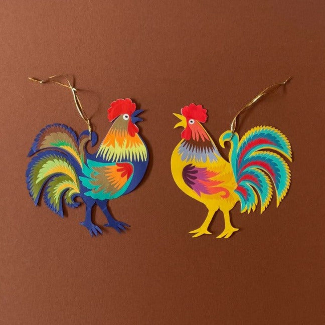 Two colourful paper cockerels decorations on a brown background.