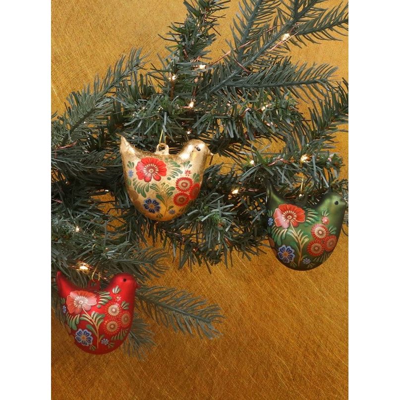Three birds decorations hanging on a Christmas tree in a dark yellow background. The birds are gold, red and green with folk designs in them.