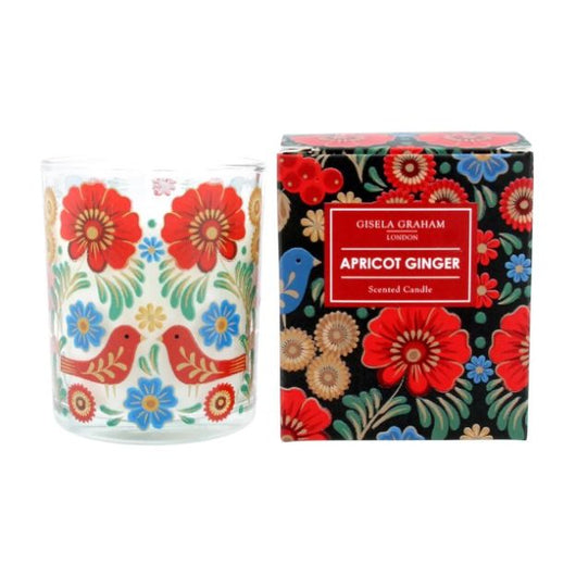 A candle in a glass container with illustrations of flowers and birds in Christmas colors, red, green, yellow and blue. Besides it there is a black box with the same illustrations in Christmas colors with a red label that reads in white font 
