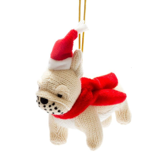 A knitted Christmas ornament shaped like a cream-colored French Bulldog, wearing a red and white Santa hat and a red scarf, hanging from a gold string against a white background.