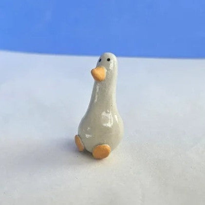 A small ceramic goose figurine with a glossy off-white body, orange beak and feet, standing on a white surface with a blue background.