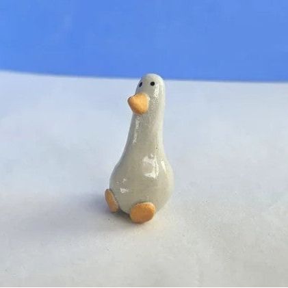A small ceramic goose figurine with a glossy off-white body, orange beak and feet, standing on a white surface with a blue background.