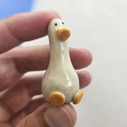 A small ceramic goose figurine with a glossy off-white body and orange beak and feet, held between fingers to show its size.