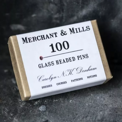 A small kraft paper box with a white label reading 'MERCHANT & MILLS 100 GLASS HEADED PINS' and 'Carlyn N.H.