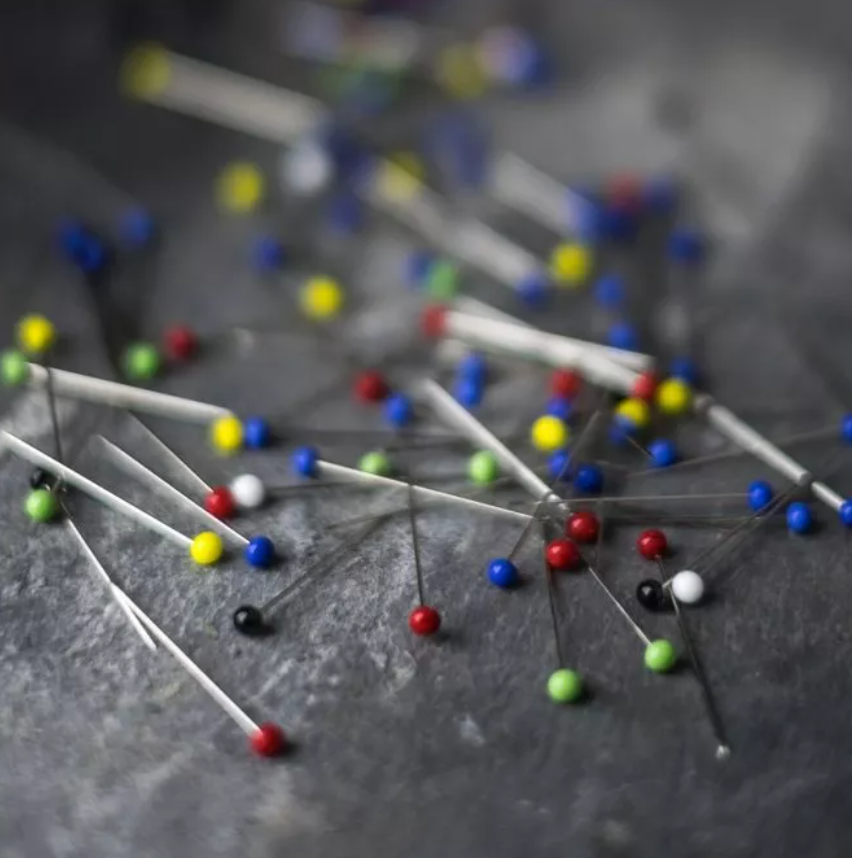 A close-up, shallow depth-of-field shot of colorful sewing pins scattered on a dark gray textured surface.