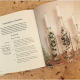 An open book on a wooden table. On the left page the instructions to make a Centre piece Classics can be read. On the left page a photograph of three glass bottles with plants inside and candles on top sit on a wooden table.