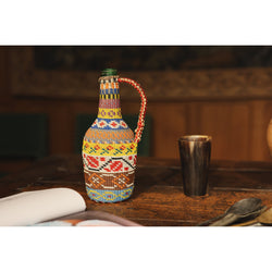 A glass bottle covered in recycled wire in colorful patterns. The bottle sits on a dark wooden table and in the background  an old tapestry on wooden walls can be seen. On the wooden table next to the bottle there are old utensils.