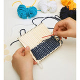 A photograph of the hands of a person crafting a coaster with navy blue loom on a white background. Besides on the left top corner there are blue, white, yellow and black yarns.