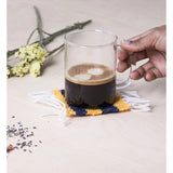 A photograph of a hand placing a glass coffee mug on a yellow and blue coaster. On the right top corner there are a few white flowers.