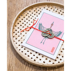 Lucky post card set with the front one showing an illustration of a bird. The postcards are tied with a red thread and three copper medals. The luck card sets are on top of a wooden braided circular plate.
