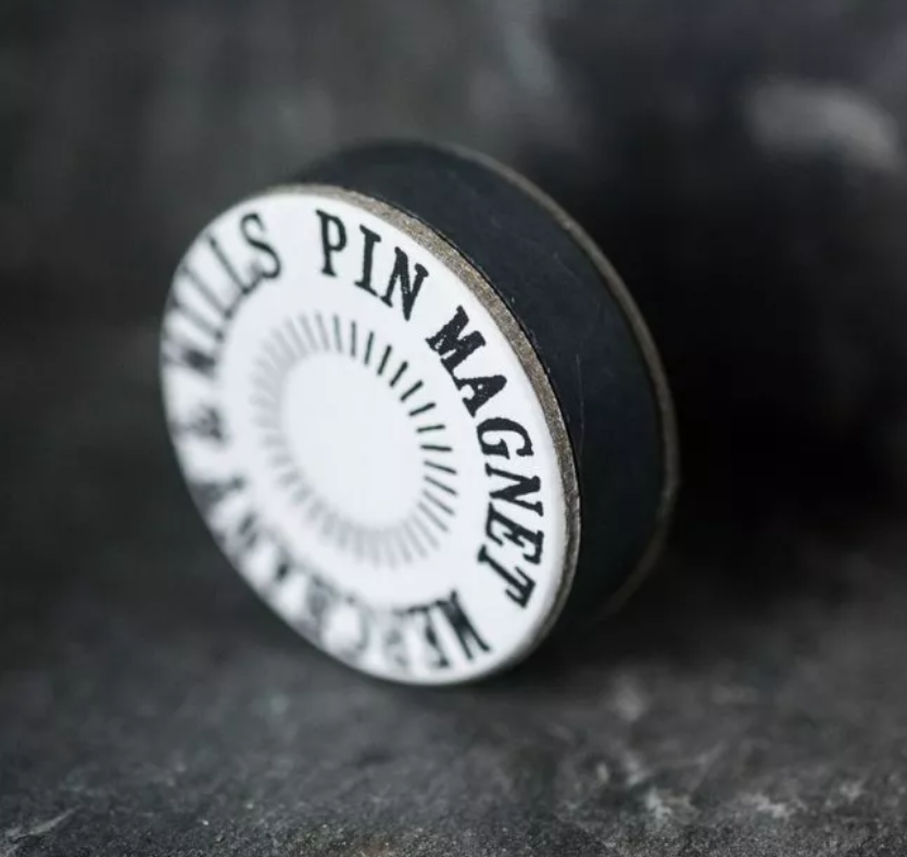 A circular black and white pin magnet tool with the text 'KILLS PIN MAGNET' printed on it, resting on a dark textured surface.