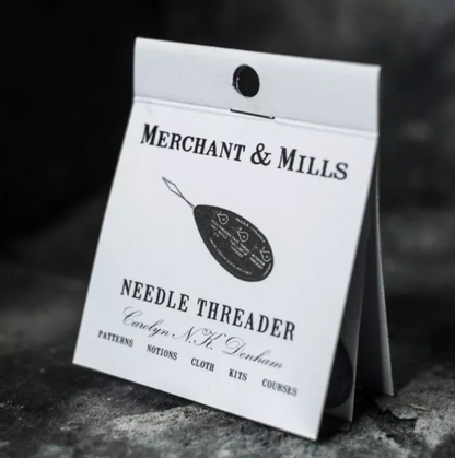 A packaged needle threader by Merchant & Mills, shown in a white hanging card package on a dark textured surface. The package features black text and an illustration of a black thread spool with a needle threaded through it.