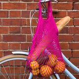 A pink cotton bag with oranges and bread inside hanging from a bike handle.