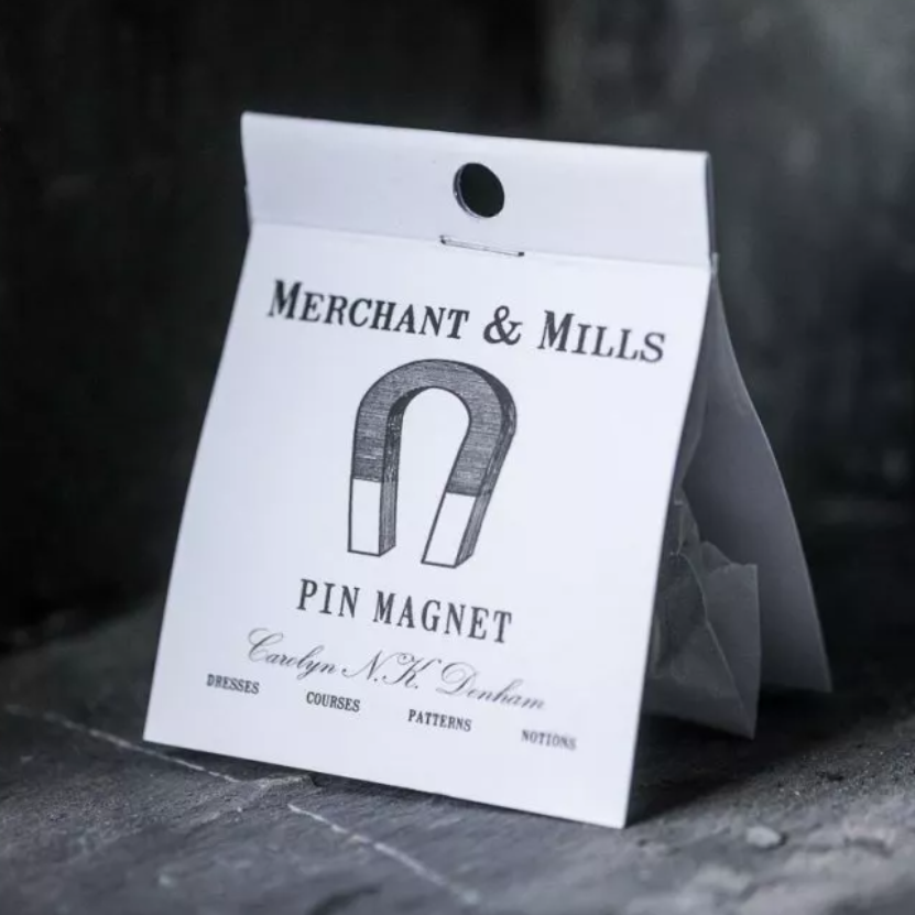 A small white paper card package for a Merchant & Mills pin magnet, photographed on a dark textured surface. The card shows a simple line drawing of a horseshoe magnet and the text 'MERCHANT & MILLS' and 'PIN MAGNET'.