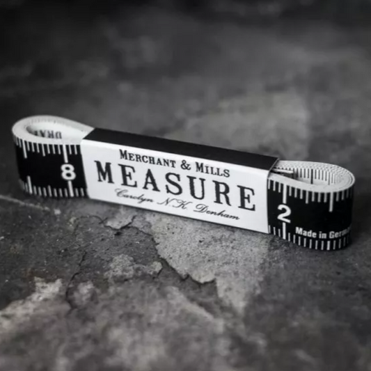 A vintage-style measuring tape coiled horizontally on a textured dark gray surface. The tape is black and white with printed measurement markings and a central paper band reading 'MERCHANT & MILLS MEASURE' and 'Carlsberg N.