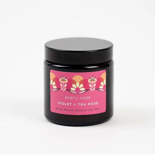 A Violet and Tea Rose Candle on a white background. The container is black glass with a plastic black lid. The label is pink with illustrations of roses in shades of pink, orange and cream details.  In small black font it reads 