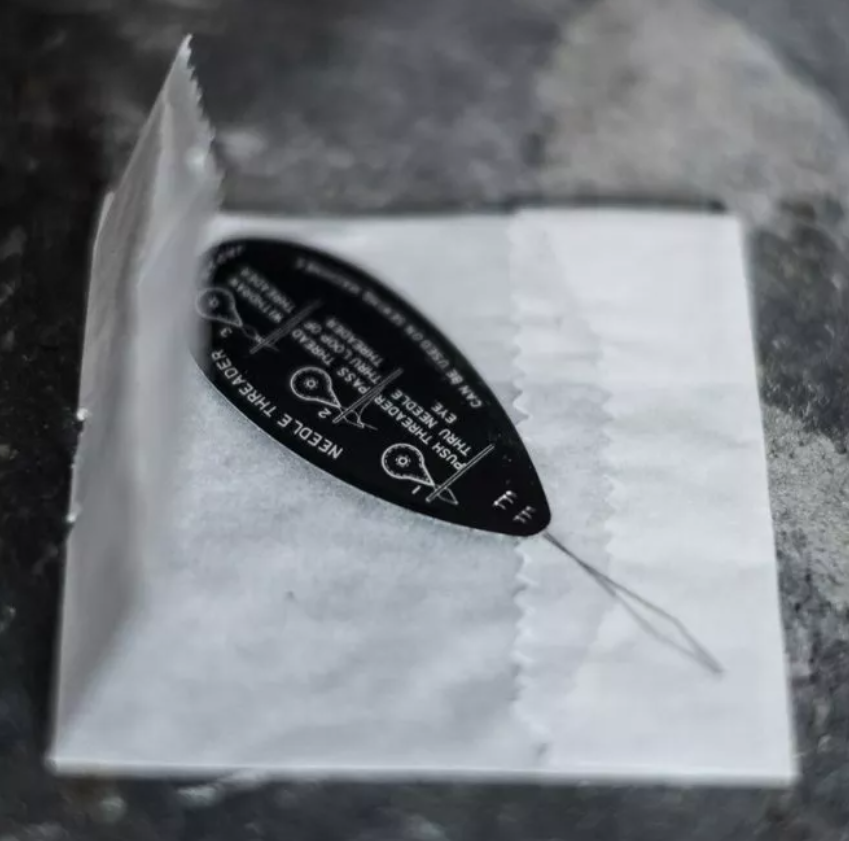 A black teardrop-shaped sewing needle with white printed instructions and text, resting on a white textured paper pad inside a clear plastic container.