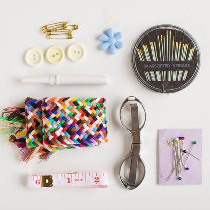 A flat-lay arrangement of sewing and crafting tools on a white background, including a multicolor braided cord, three cream buttons, gold safety pins, a blue flower embellishment, a round tin of assorted needles, a white stitch marker, a metal ring binder clip, a pink measuring tape, and pins on a lavender card.