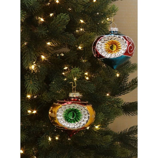 Two vintage dimpled baubles hanging on a pine tree with fairy lights on a beige background. The bauble are red and blue with details in green and gold.