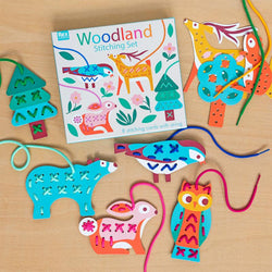 A stitching set scattered on a wooden background. The cards feature a blue bear, a tree, a pink rabbit, an orange and blue owl, a blue and red bird, a pine tree, an orange squirrel and a yellow deer.