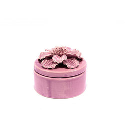 A round, pale pink ceramic trinket box with a lid on a plain white background. On the lid sits an ornate ceramic carved flower with petals and stems.